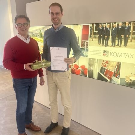 Zwei Männer mit Urkunde und Geschenk vor KOMTAX-Wand