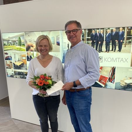 Frau mit Blumenstrauß und Mann mit Umschlag vor KOMTAX-Wand
